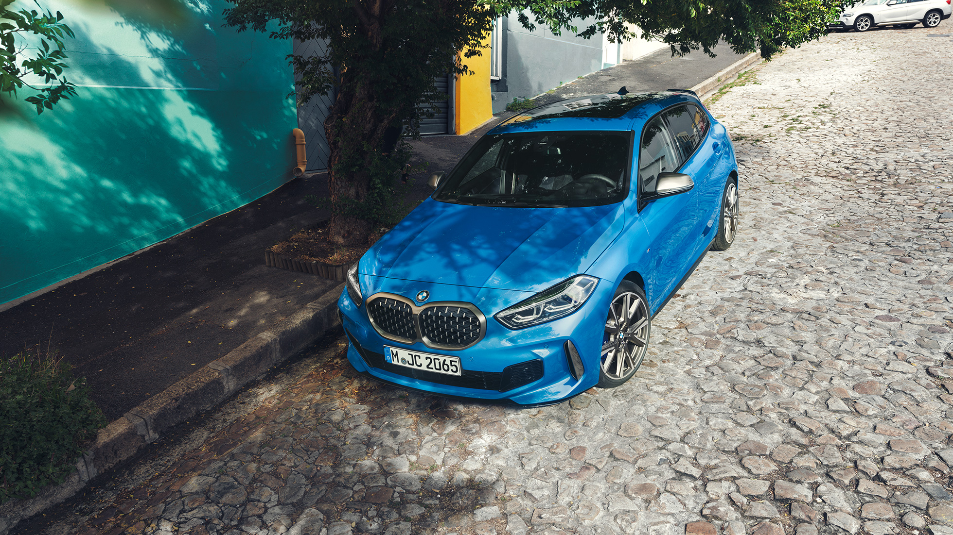 BMW 1 Serie langs de kant van de weg. BMW 1 Serie in Misano Blau metallic, driekwart vooraanzicht, langs de weg geparkeerd en van bovenaf gezien.