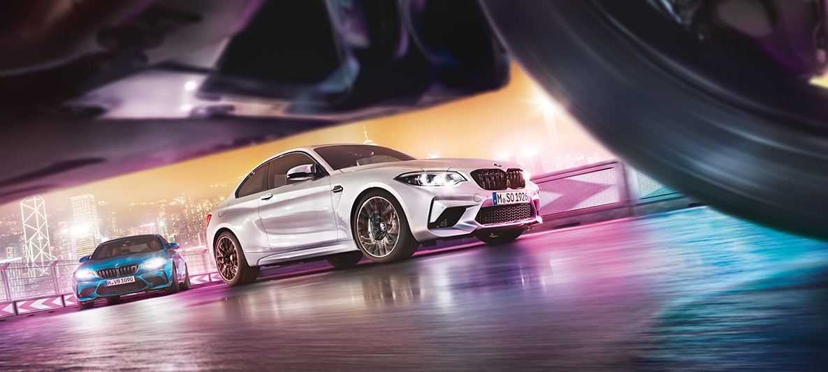 BMW M2 Competition op spiegelend asfalt Twee BMW’s M2 Competition op het circuit met op de achtergrond een nachtelijke skyline, gezien vanaf de grond.