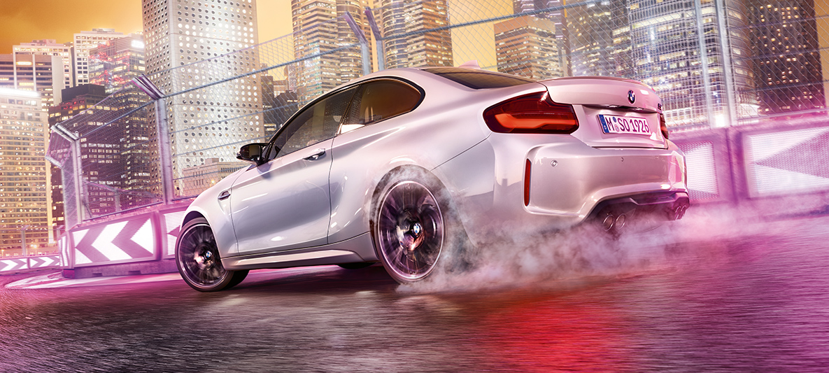 BMW M2 Competition geeft gas Witte BMW M2 Competition driftend op het circuit met op de achtergrond een nachtelijke skyline.