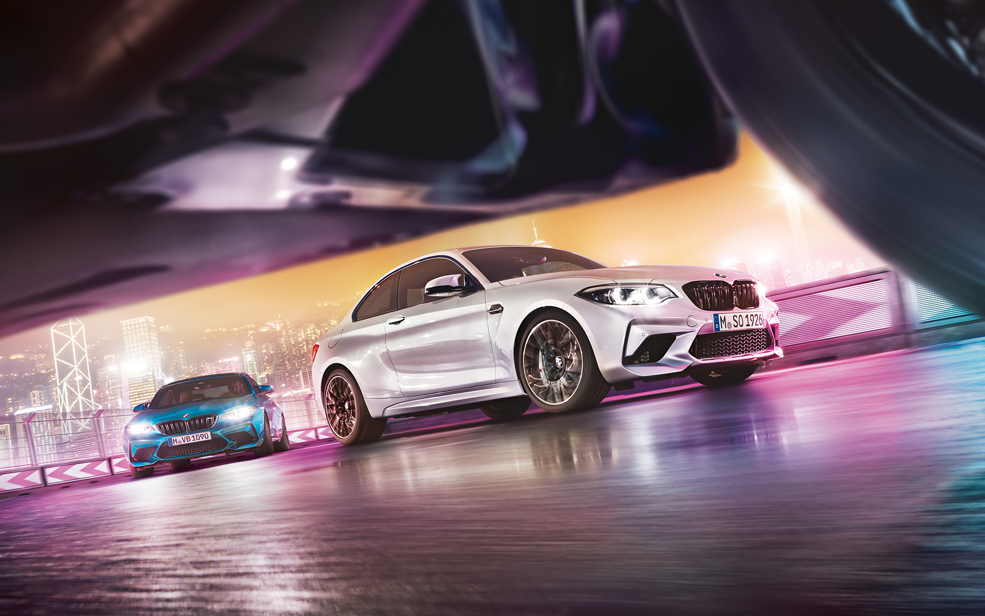 BMW M2 Competition op spiegelend asfalt Twee BMW’s M2 Competition op het circuit met op de achtergrond een nachtelijke skyline, gezien vanaf de grond.