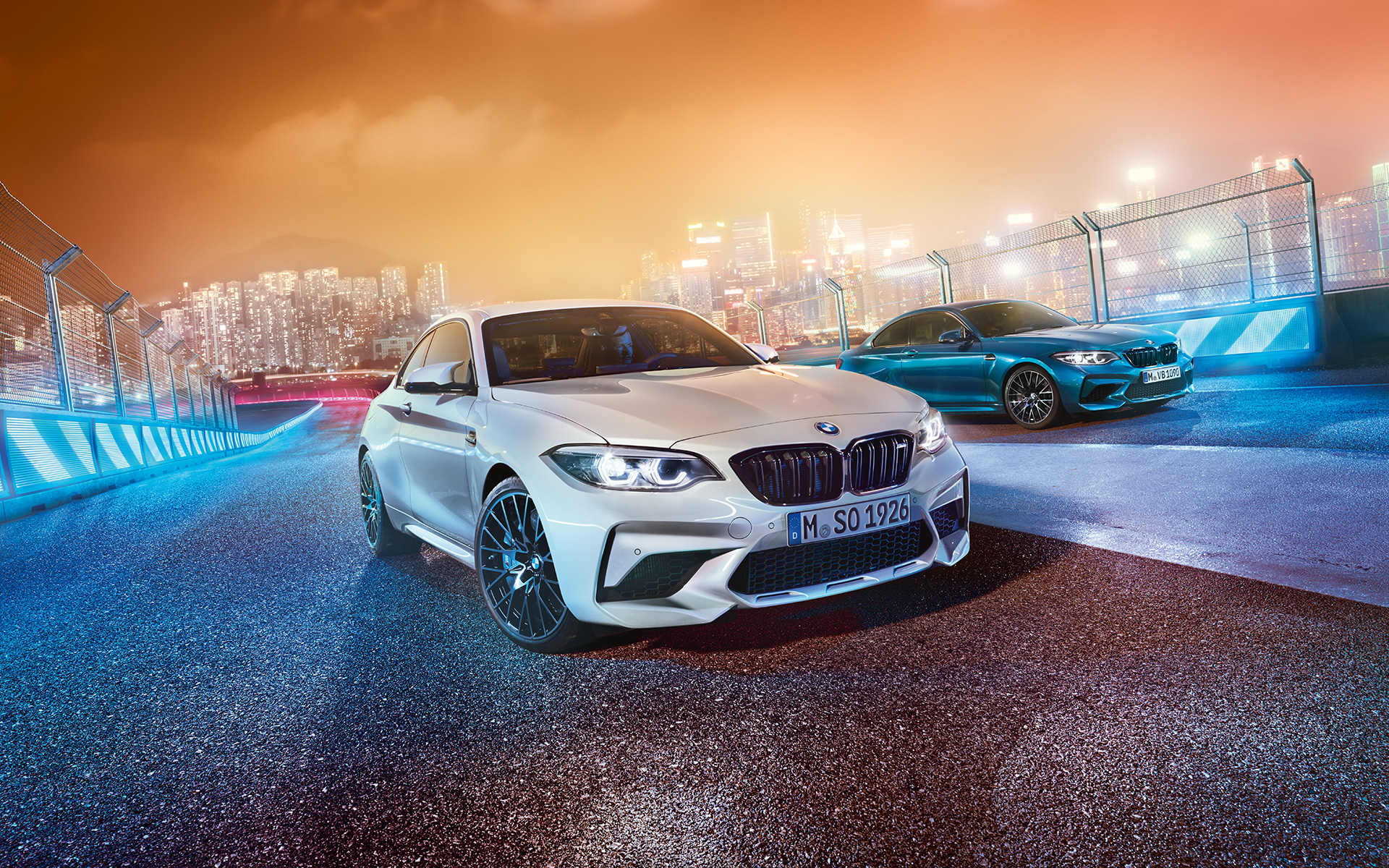 Twee BMW’s M2 Competition op het circuit Witte en blauwe stilstaande BMW M2 Competition schuin van voren gezien met op de achtergrond een nachtelijke skyline.