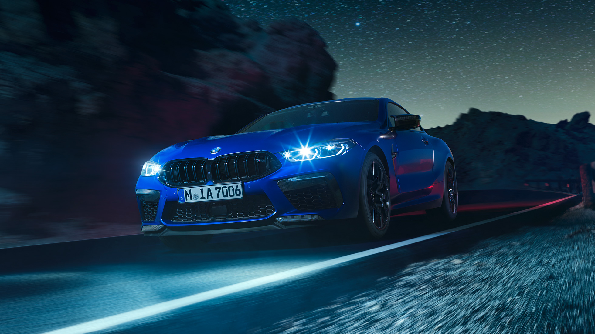 Rijdende BMW M8 Competition Coupé, driekwart vooraanzicht. BMW M8 Competition Coupé in Marina Bay Blau metallic met LED koplampen, driekwart vooraanzicht nacht.