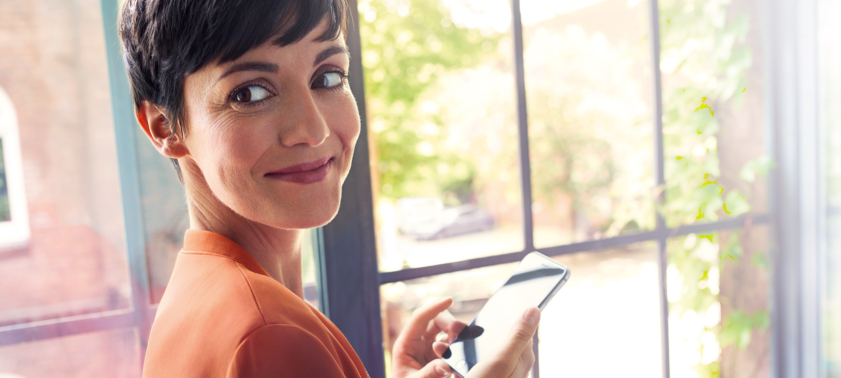 Donkerharige vrouw met smartphone Donkerharige vrouw in oranje jas staat met haar smartphone bij het raam en glimlacht