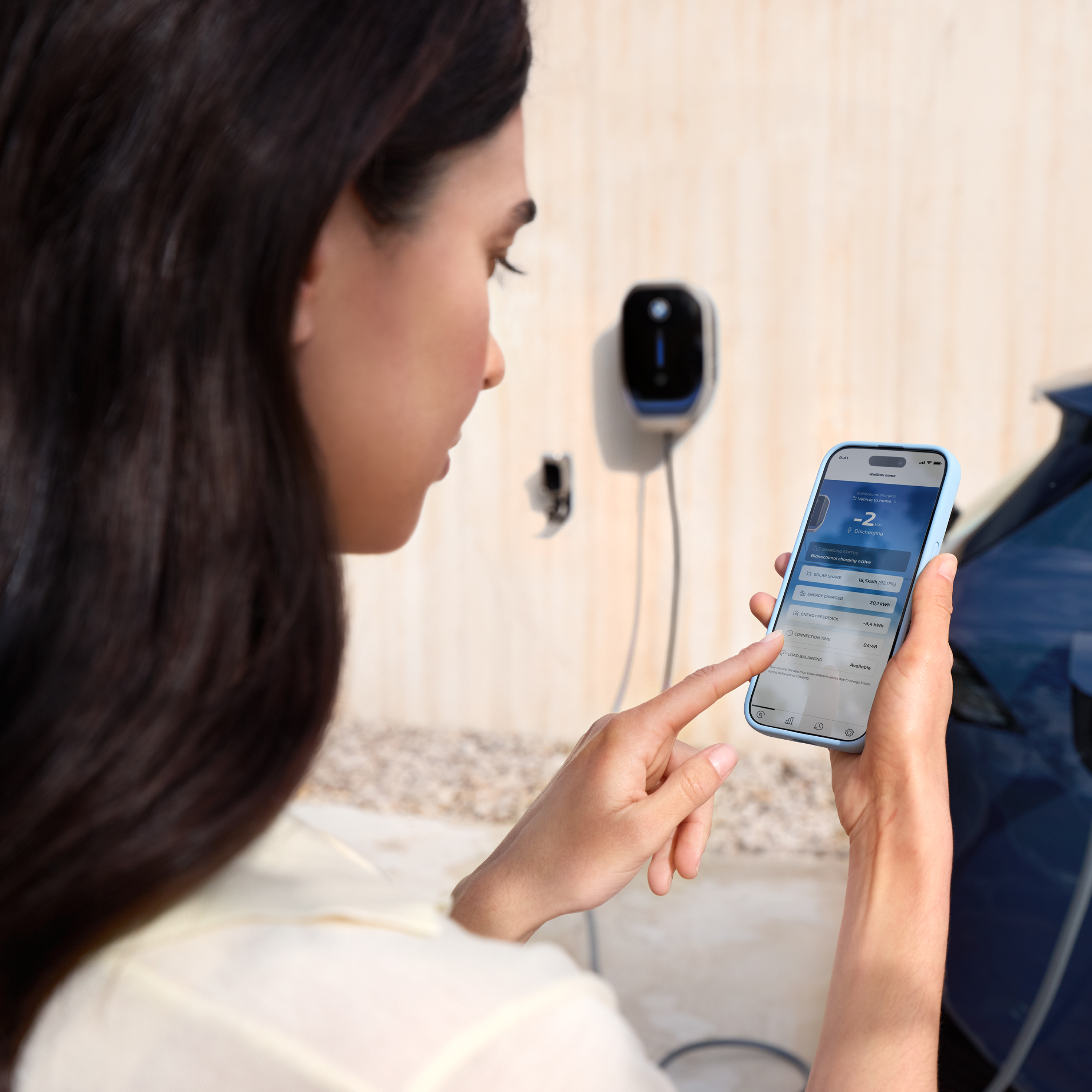 Een vrouw kijkt naar haar smartphone met een BMW wallbox op de achtergrond